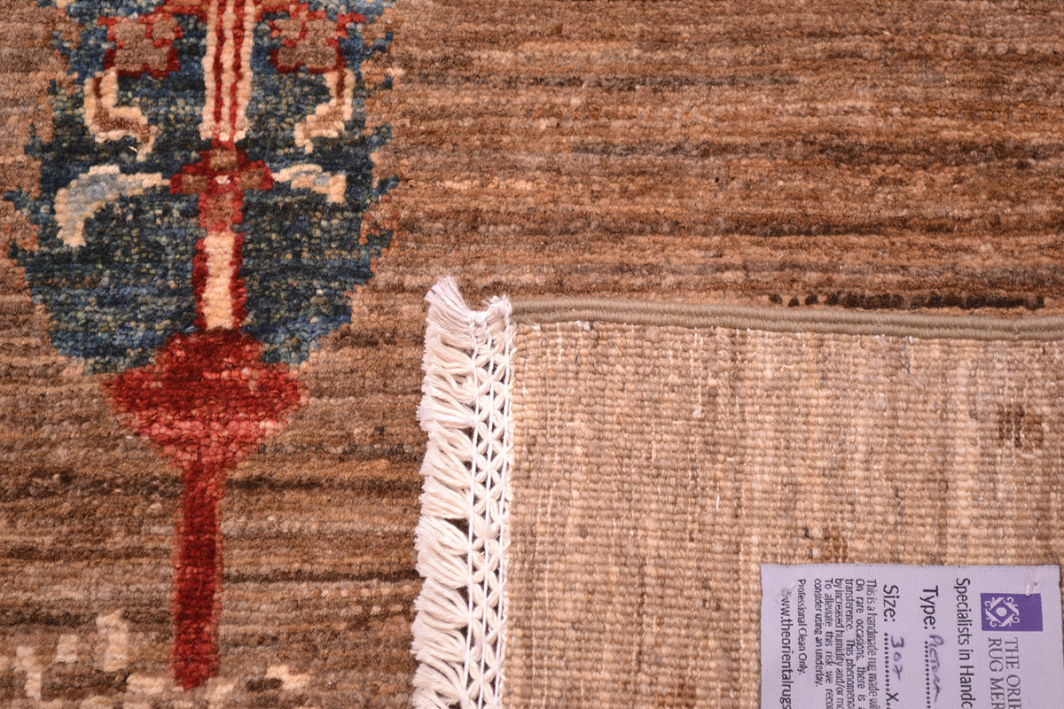 Close-up of a corner turned up showing handknotting weave of a Pictorial Afghan Loribaft carpet showing a tan and brown textured base, a partial blue and red decorative motif, white fringe, and a product label.
