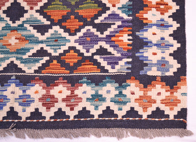 A close-up of a corner of colourful Kilim handwoven rug showing its geometric pattern and fringed edge. The design features a dark navy blue background with cream, orange, teal, purple, and red diamond and star motifs.