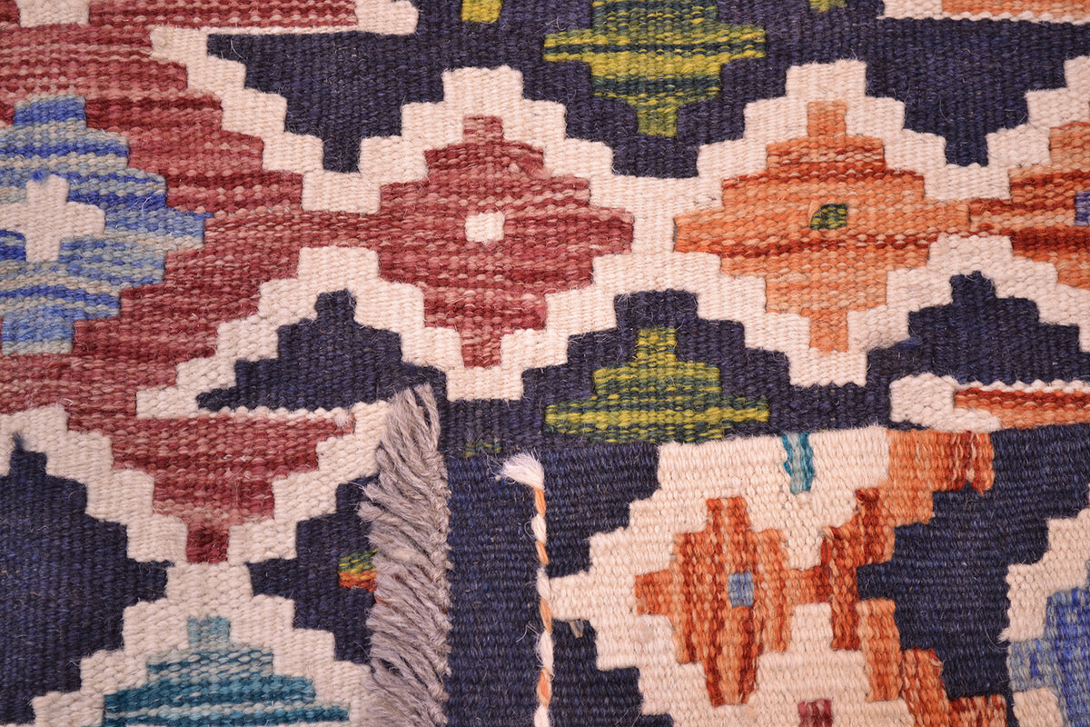 Close-up of a corner turned up showing the handwoven weave of a colourful Afghan Maimana kilim rug showing a geometric pattern with navy blue, terracotta red, orange, cream, and light blue threads, featuring a fringed edge.