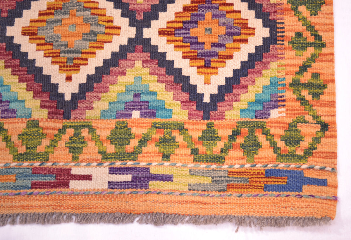 A close-up of a corner of a handwoven multicolour Kilim short runner showing its geometric diamond and zigzag patterns in orange, purple, green, blue, and cream, with a fringed edge visible at the bottom.