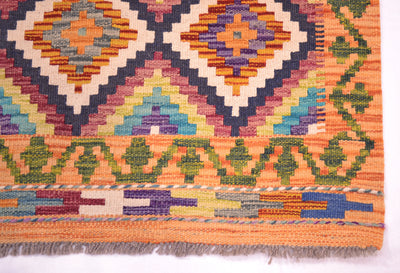 A close-up of a corner of a handwoven multicolour Kilim short runner showing its geometric diamond and zigzag patterns in orange, purple, green, blue, and cream, with a fringed edge visible at the bottom.