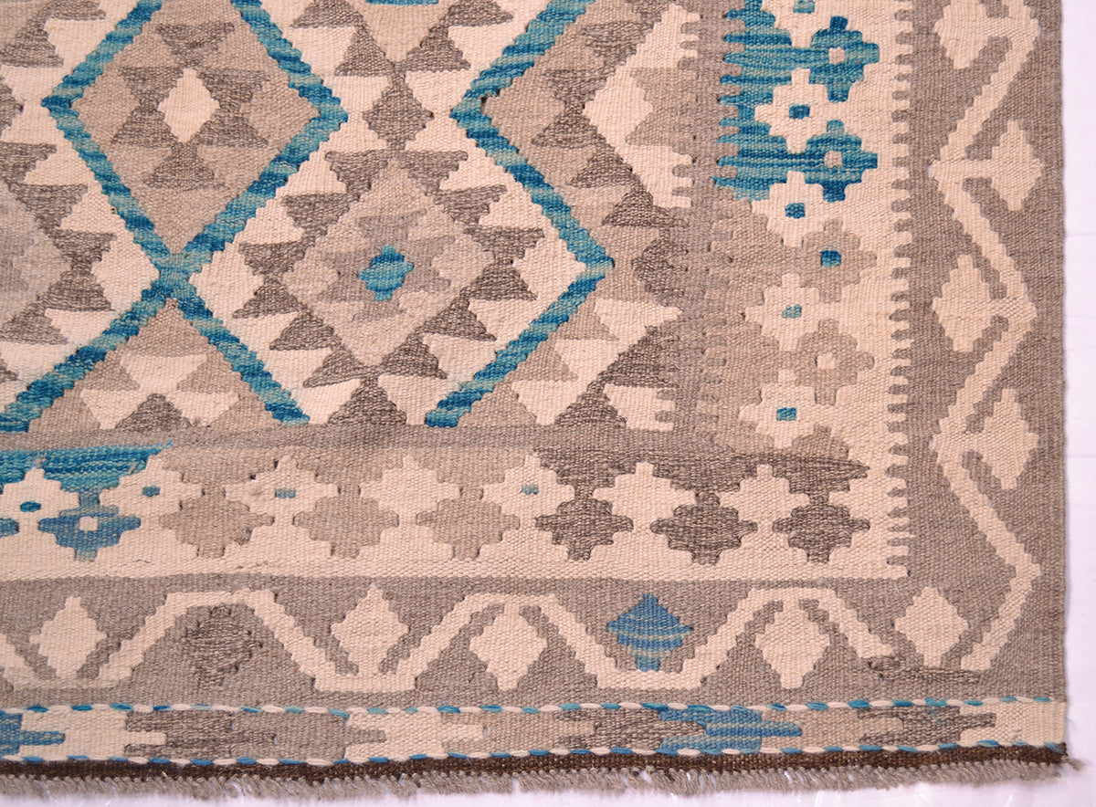 A close-up of a handwoven large Afghan Kilim rug showing a geometric pattern in beige, grey, and turquoise blue, with a fringed edge visible at the bottom.