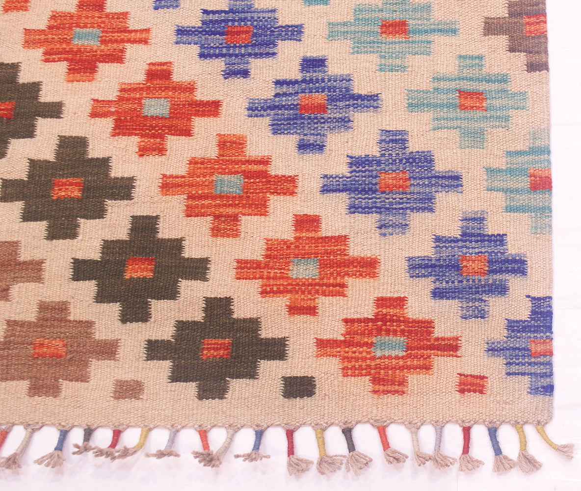 A close-up of a corner of a handwoven Afghan kilim hallway runner rug with a geometric pattern in red, blue, brown, and light blue on a beige background, finished with a multicolour tassel fringe.
