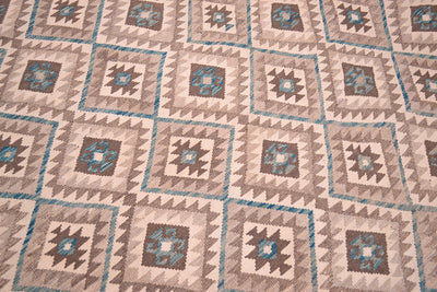 A close-up of a woven large Kilim rug featuring a repeating geometric pattern in beige, brown, teal, and cream.