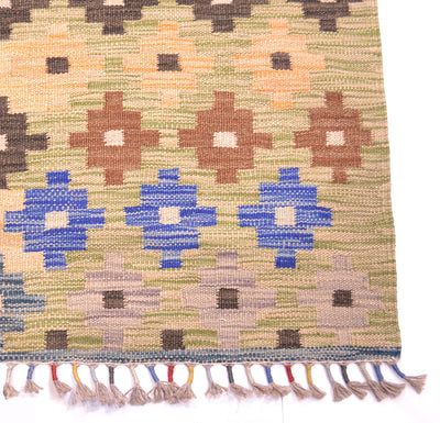 A close-up of a woven kilim runner rug showing its detailed pattern and fringed edge. The rug features a multicolour design with blue, brown, beige, and green geometric motifs on a textured woven background.