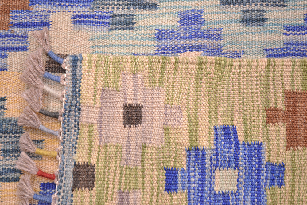 Close-up of the weave of a handwoven multicolour long Kilim runner rug corner showing a cream base with blue, green, and tan geometric motifs and fringed tassels along one edge.
