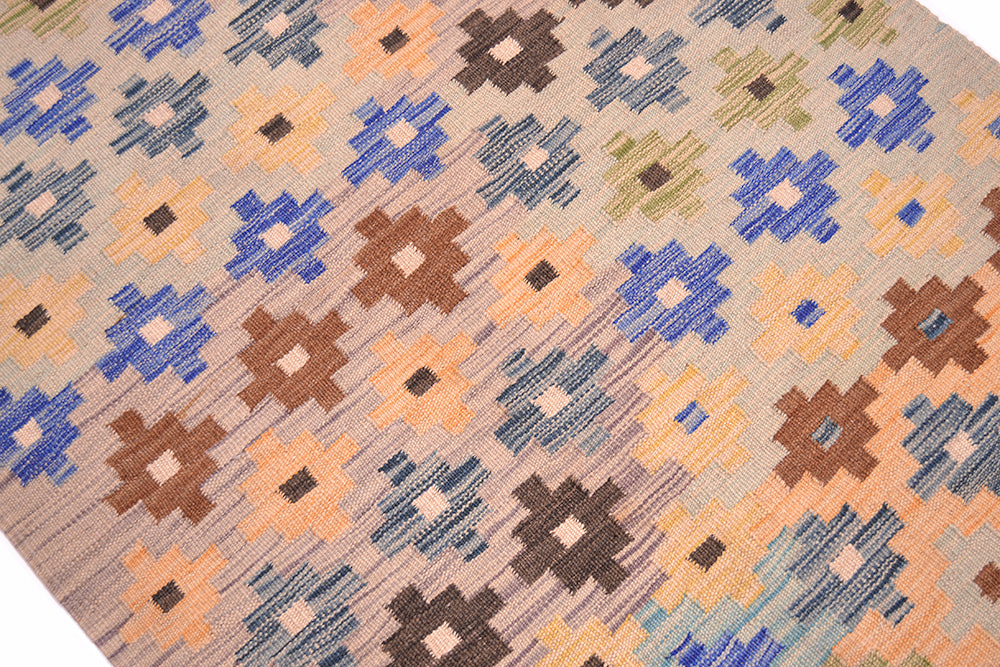 A close-up of a handwoven Afghan Kilim runner with a geometric pattern in blue, brown, peach, and olive on a beige background.