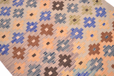 A close-up of a handwoven Afghan Kilim runner with a geometric pattern in blue, brown, peach, and olive on a beige background.