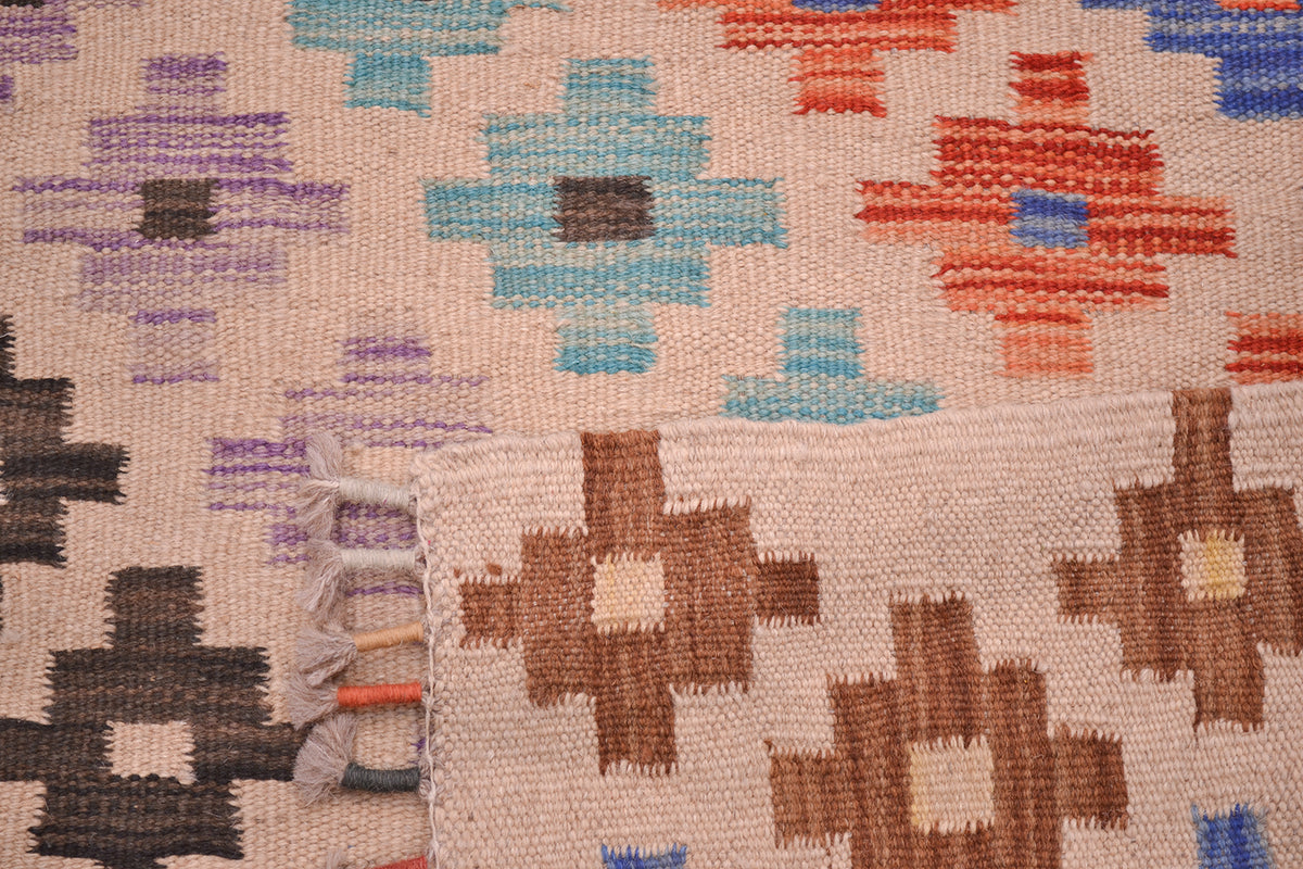 Close-up of the weave of a handwoven multicolour Afghan Kilim runner rug with a geometric ziggurat pattern in beige, brown, orange, blue, purple, and black, showing the texture and fringe of one corner.