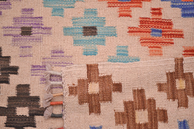 Close-up of the weave of a handwoven multicolour Afghan Kilim runner rug with a geometric ziggurat pattern in beige, brown, orange, blue, purple, and black, showing the texture and fringe of one corner.