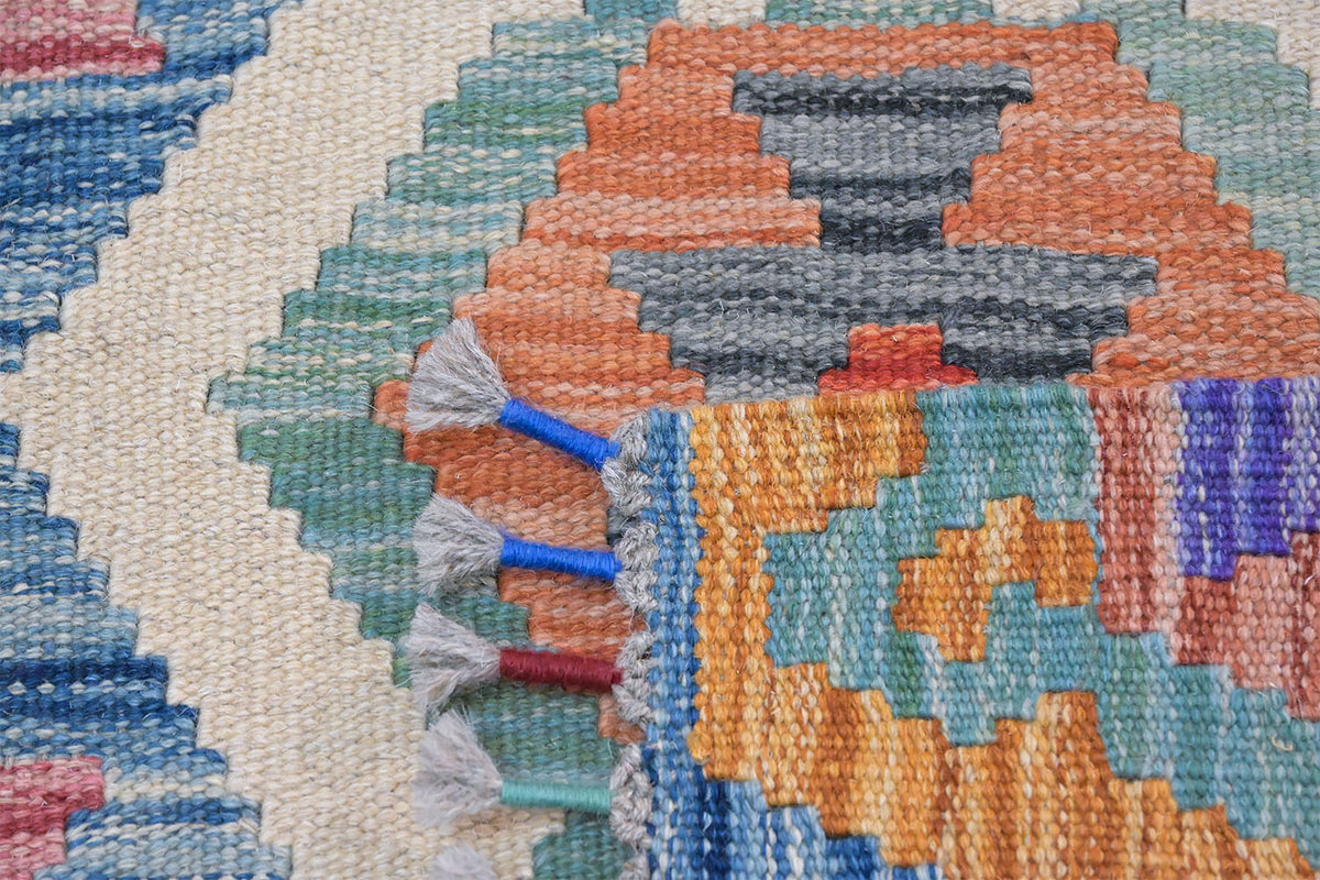 Close-up of a woven rug with a multicolor geometric pattern, featuring orange, blue, green, gray, and cream sections with white tassels on the edge.