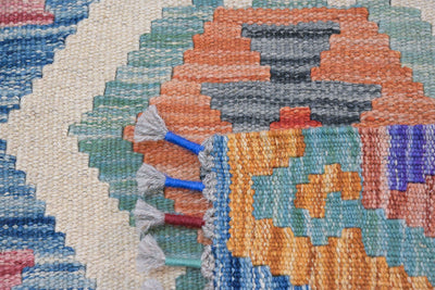 Close-up of a woven rug with a multicolor geometric pattern, featuring orange, blue, green, gray, and cream sections with white tassels on the edge.
