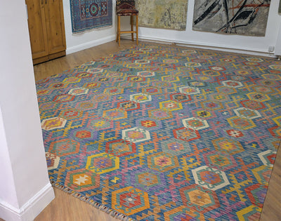 A Giant Handwoven Afghan Kilim rug with a colourful geometric pattern, shown in a room with wooden floors and other wall hangings.