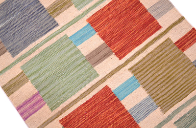 A close-up of a woven Modern Kilim short runner with a geometric contemporary pattern in muted tones of red, green, blue, beige, brown, and purple on a light cream background.