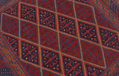 A close-up of a woven Mushwani square wool rug with a geometric diamond pattern in deep red, navy blue, and tan.