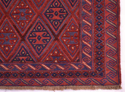 A close-up of a patterned square Moshvani rug corner showing geometric diamond motifs in red, navy blue, and beige, with a fringed edge and woven texture.