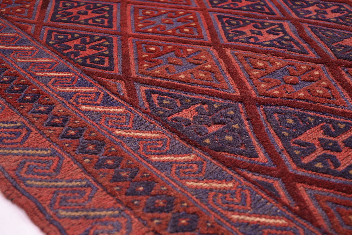 A close-up of an Afghan handwoven Moshvani Kilim rug with an intricate geometric pattern in rust red, dark navy blue, and brown.