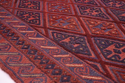 A close-up of an Afghan handwoven Moshvani Kilim rug with an intricate geometric pattern in rust red, dark navy blue, and brown.