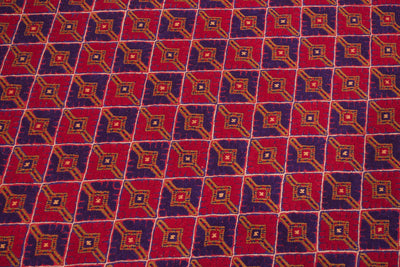 A close-up of a fine Mushwani rug / carpet with a repeating geometric diamond pattern in red, purple, orange, and cream.
