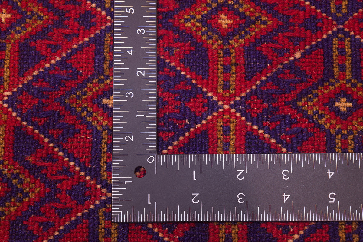 A close-up of a fine Afghan Mushwani carpet with a dark metal ruler placed over it to show scale. The fabric has a geometric tribal-style pattern in red, purple, and gold.