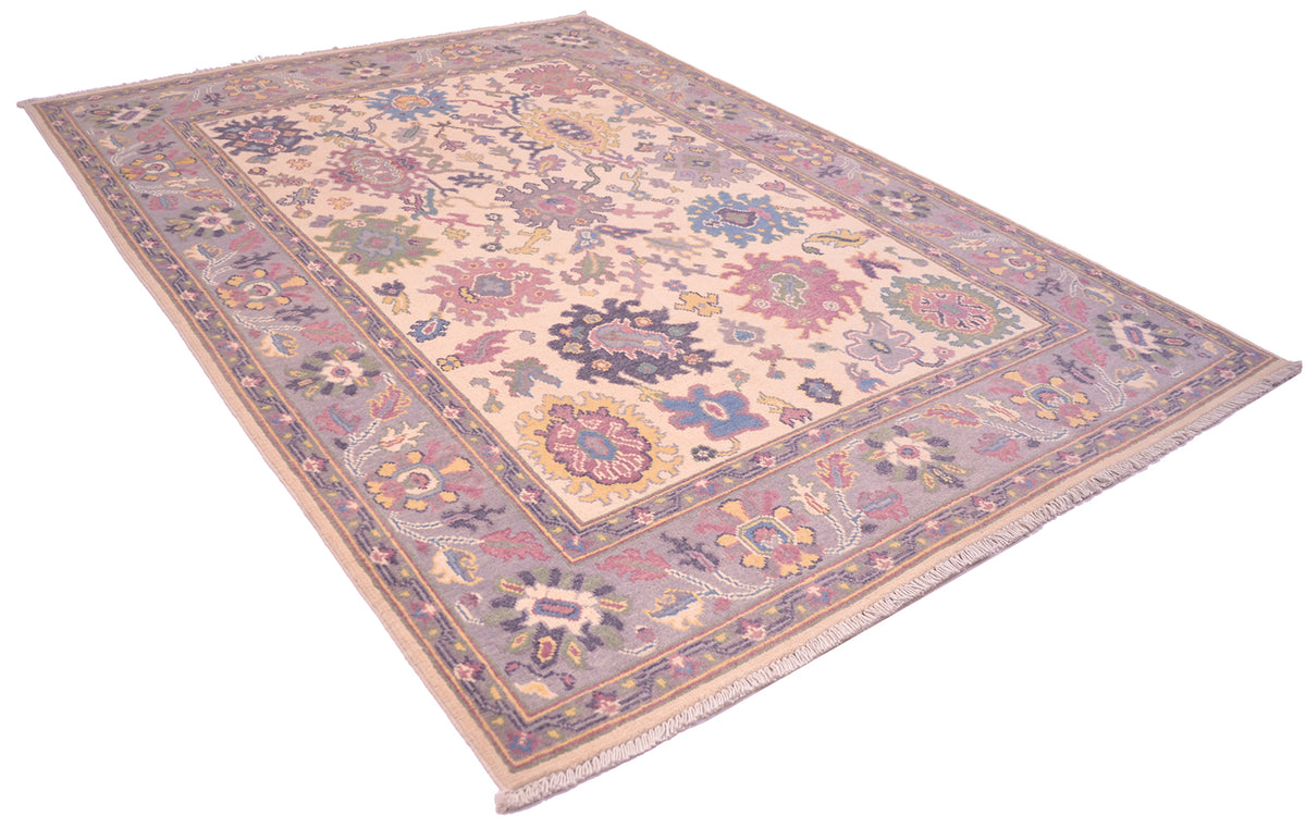 Handmade Oushak rug angled view, cream field with mauve, blue, and gold floral border.
