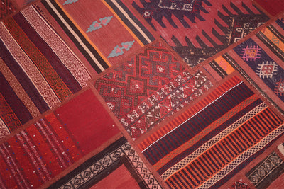 A close-up of a patchwork kilim rug made of multiple red, rust, orange, brown, and black patterned sections with geometric and striped motifs.