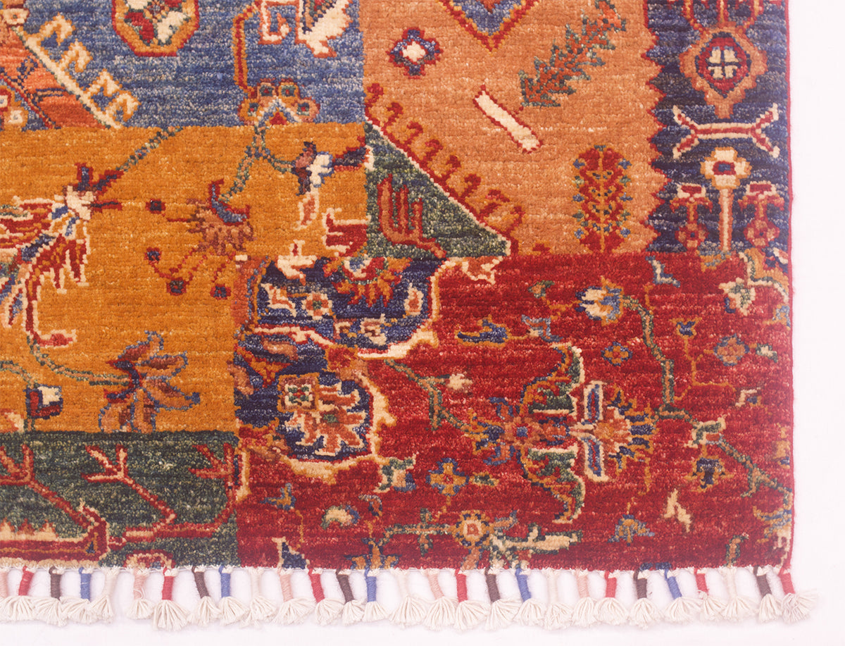 A close-up of a patterned area rug showing its corner and fringe. The rug features a patchwork design with multiple colors including orange, red, blue, green, and cream, with a white fringed edge at the bottom.