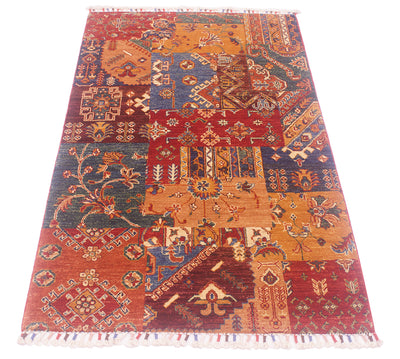 A rectangular Afghan patchwork-style rug with a traditional geometric and floral pattern, featuring multiple panels in rust red, terracotta, navy blue, ochre orange, and cream, finished with white fringe on the ends.