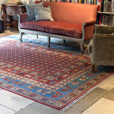 A large traditional style handmade Afghan Pazyryk carpet with a red and blue patterned design, styled in a cozy living room setting under a coral velvet sofa.