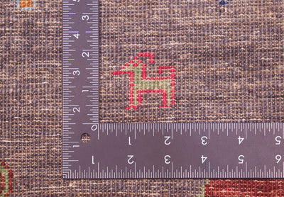 Close-up of a hand-knotted Afghan Loribaft pictorial runner with a small red and green embroidered motif on a brown textured background, with a metal L-square ruler placed at the top left to show scale of the knotting.