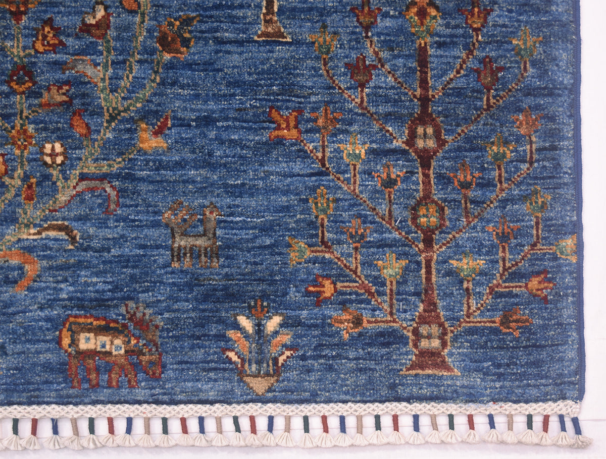A close-up of a corner of a pictorial blue Loribaft rug with woven botanical and animal motifs, including a tree, birds, and a small camel, finished with a white braided fringe at the bottom.