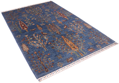 A rectangular pictorial blue Loribaft rug with a blue background and an intricate, traditional tribal-style pattern featuring trees, birds, and geometric shapes in brown, beige, and muted orange. The rug has a white fringed border on one end.
