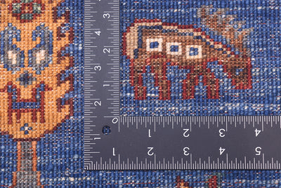 Close-up of the hand-knotted back of a pictorial blue Loribaft Rug showing a blue field with a brown animal motif and part of an orange decorative figure, overlaid with a dark ruler to show scale of the knot density.