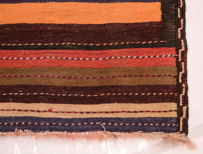 A close-up of a striped woven vintage Kilim  runner rug corner showing horizontal bands in orange, dark brown, rust red, olive green, beige, and navy blue with fringed edges.