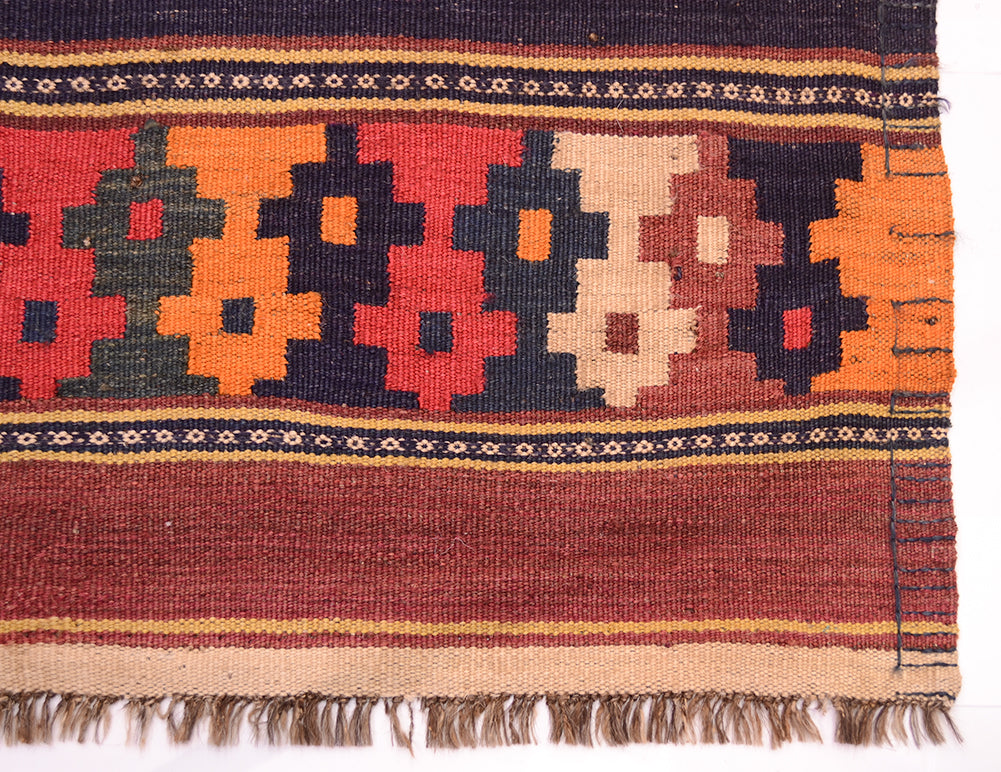 A close-up of a handwoven old Afghan kilim rug showing its geometric pattern and fringe. The rug features a multi-coloured design with red, orange, beige, and dark brown shapes, bordered by striped bands of beige, brown, and black, and finished with a fringed edge.