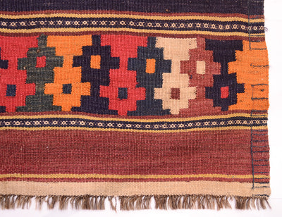 A close-up of a handwoven old Afghan kilim rug showing its geometric pattern and fringe. The rug features a multi-coloured design with red, orange, beige, and dark brown shapes, bordered by striped bands of beige, brown, and black, and finished with a fringed edge.