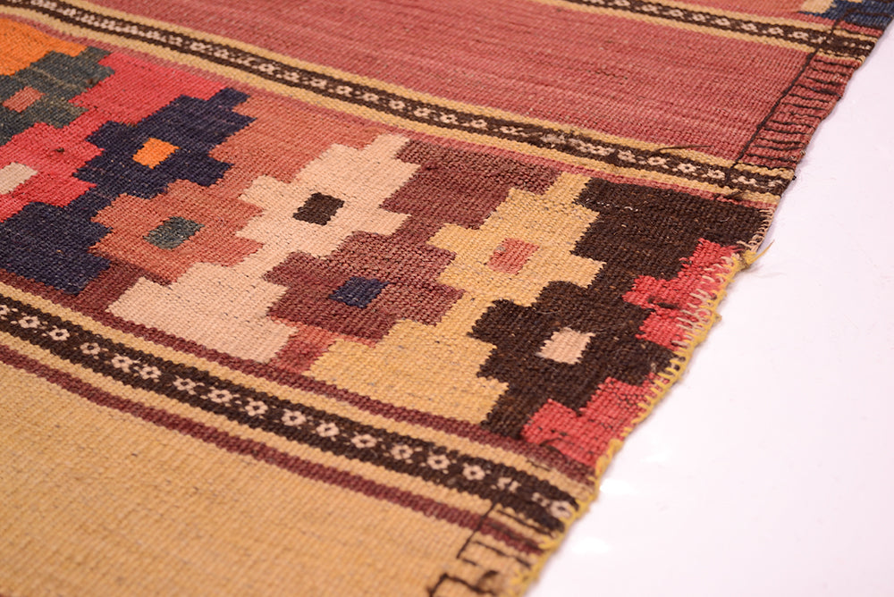 A close-up of a woven kilim rug edge showing a geometric pattern in tan, red, brown, cream, and dark navy on a white background.