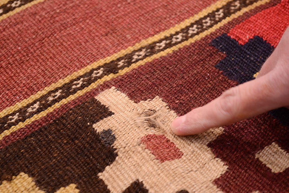 A close-up of a handwoven old Afghan Kilim rug with a person's finger pointing to a loose, frayed section of the beige pattern.
