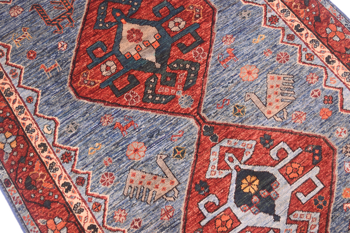 A close-up of a patterned Handmade Qashqai design rug featuring a blue field with large red geometric panels and stylised animal motifs in beige, deep teal green, and rust tones.