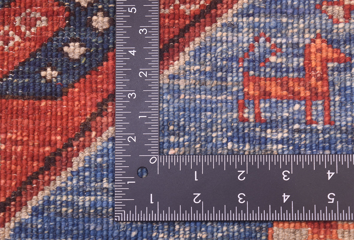 Close-up of the hand-knotted weave of a handmade Qashqai design rug showing a blue field with a red dog motif, a red-and-blue patterned section, and a dark ruler placed over the surface to indicate scale of the knot density. 
