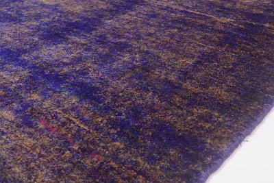 A close-up of a Handmade Sari Silk rug showing a mottled pattern of deep purple, blue, and gold tones, with a white surface visible at the bottom right corner.