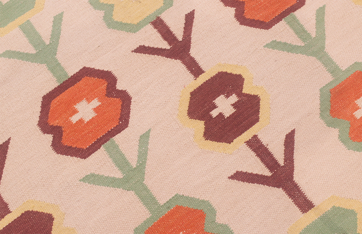 A close-up of a woven Scandinavian style Kilim rug featuring stylised floral motifs in orange, dark brown, sage green, and cream on a cream background.