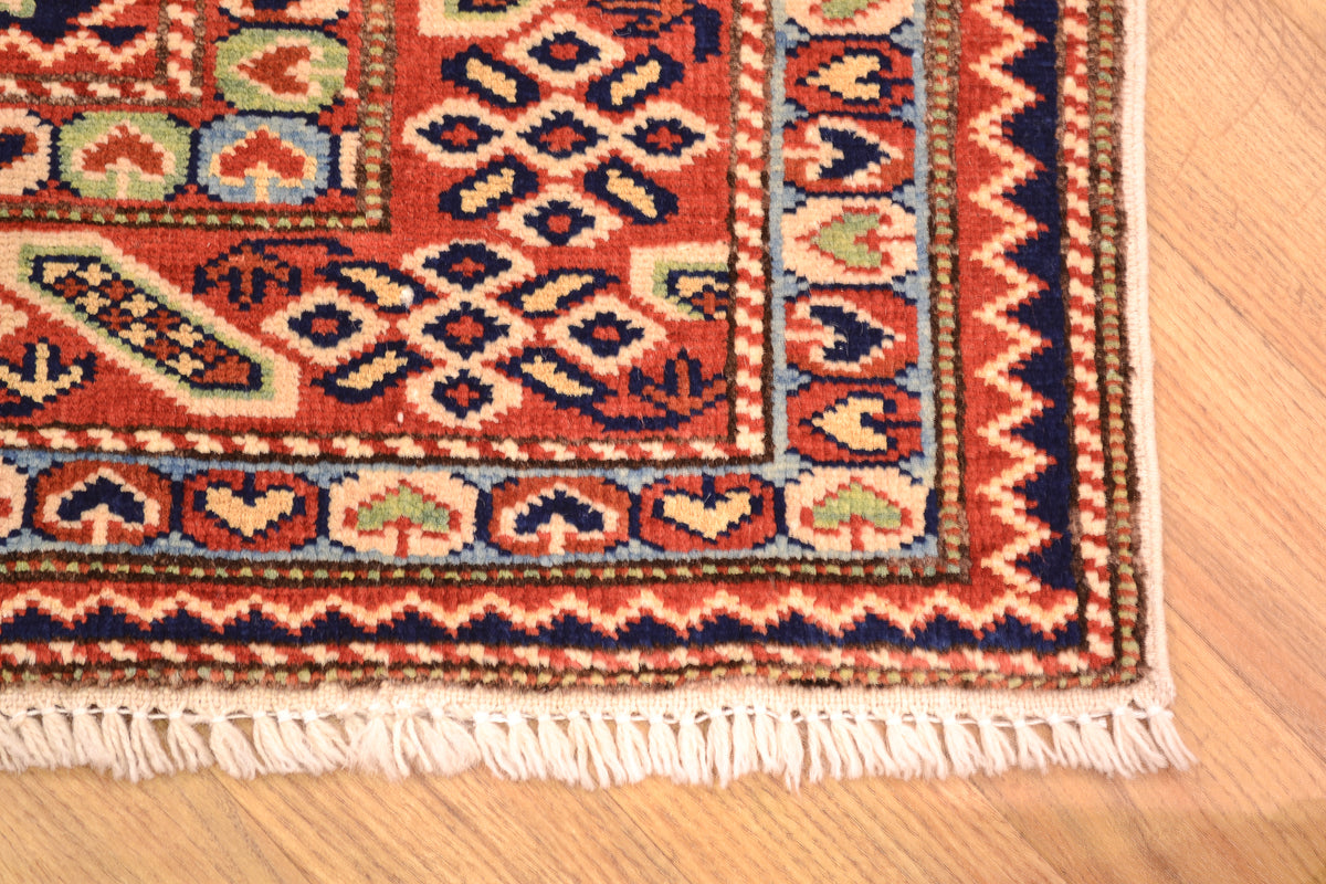 A close-up of a patterned Shirvan Afghan area rug corner on a light wood floor, showing cream fringe, red and orange field, navy and beige geometric motifs, and a blue border.