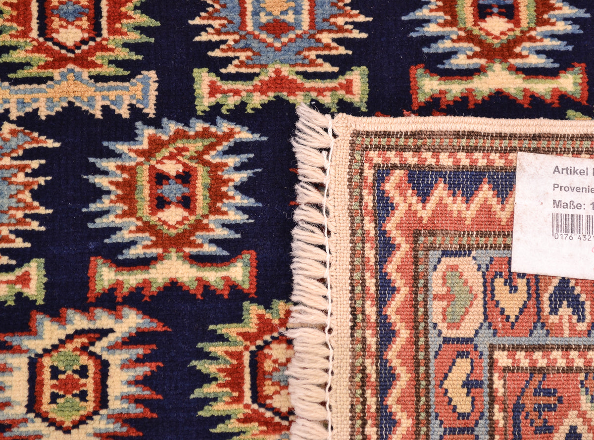 Close-up of the hand-knotted underside corner  of a patterned Afghan Shirvan rug - corner showing cream fringe, a white product tag, and detailed woven motifs in navy, rust red, beige, and light blue.