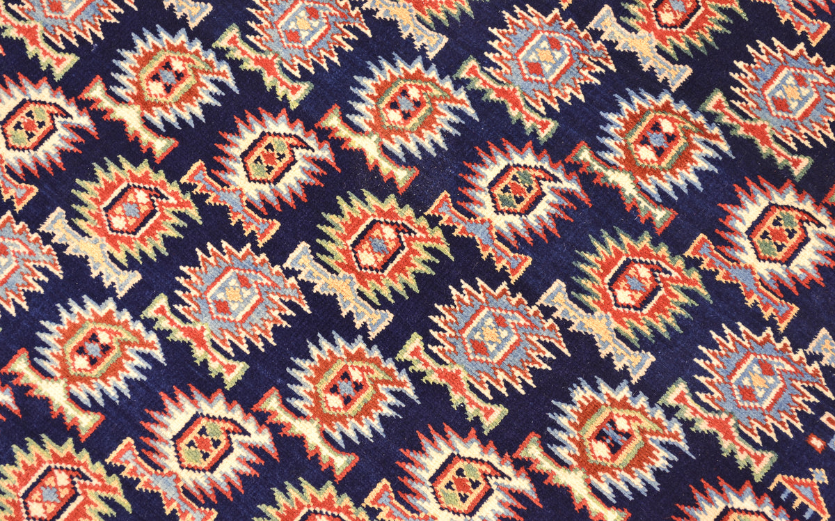 A close-up of a  fine handmade Afghan Shirvan rug with a repeating geometric pattern on a dark navy background. The pattern features orange, beige, and light blue diamond and marasali boteh motifs.