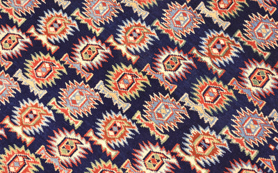 A close-up of a  fine handmade Afghan Shirvan rug with a repeating geometric pattern on a dark navy background. The pattern features orange, beige, and light blue diamond and marasali boteh motifs.