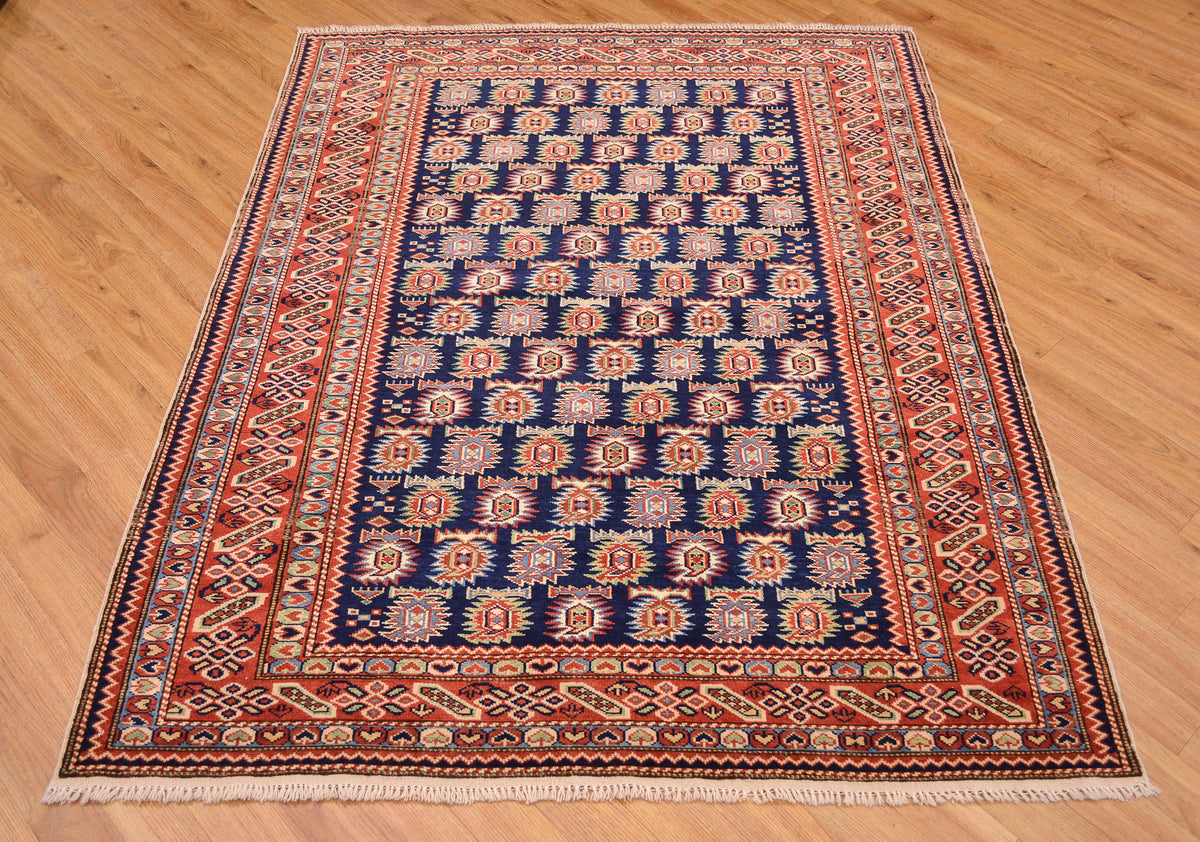 A rectangular traditional hand-knotted fine Afghan Shirvan area rug with a navy blue centre field, red and beige geometric borders, and white fringe, displayed on a light brown wooden floor.