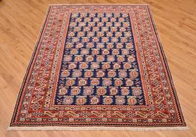 A rectangular traditional hand-knotted fine Afghan Shirvan area rug with a navy blue centre field, red and beige geometric borders, and white fringe, displayed on a light brown wooden floor.