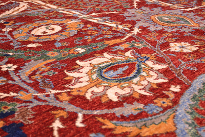 A close-up of a traditional patterned rug with a deep red field, featuring cream, orange, blue, and green floral and paisley motifs.