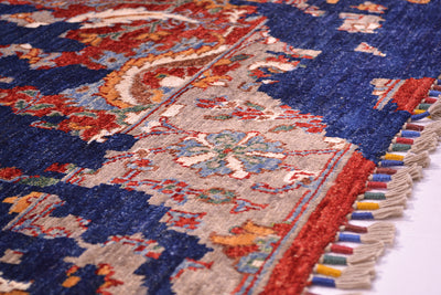 A close-up of a traditional patterned rug with a deep blue field, beige central panel, red border, and multicolored fringe tassels.